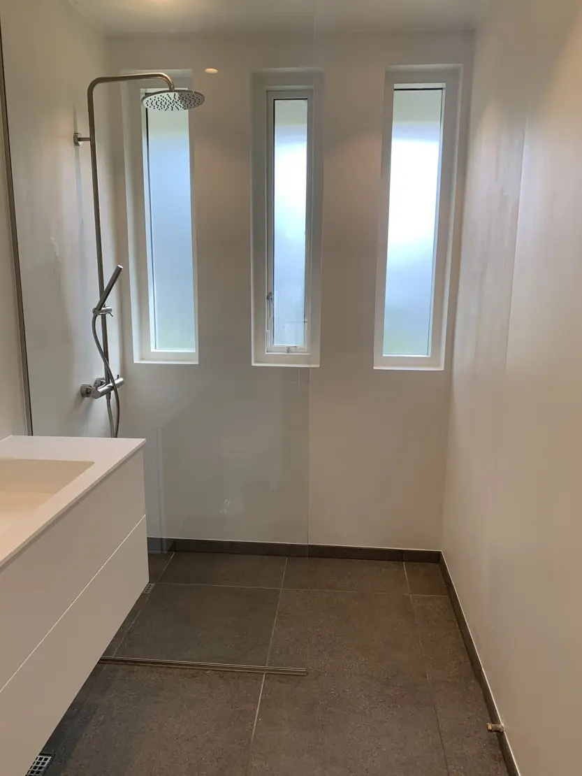 Baño de microcemento con paredes blancas, suelo de azulejos y tres pequeñas ventanas