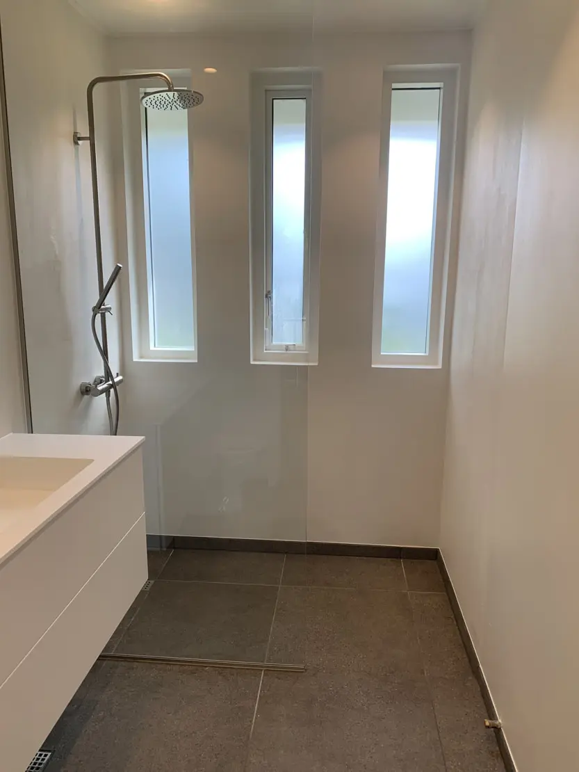 Mikrocementa vannas istaba ar baltām sienām, flīžu grīda un trīs maziem logiem